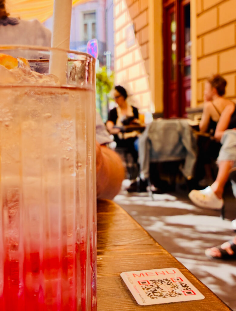 Glas mit rotem Getränk und Eiswürfeln auf Holztisch vor unscharfem Straßencafé mit mehreren sitzenden Personen