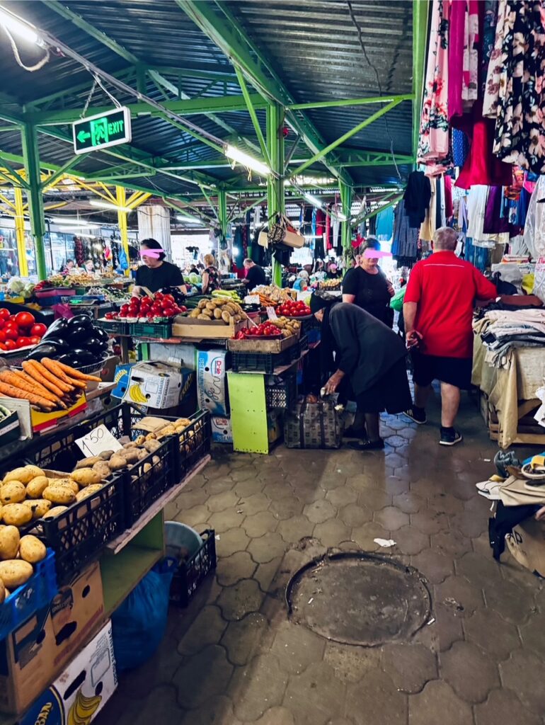 Markthalle mit Ständen für Gemüse und Kleidung, mehrere Personen beim Einkaufen und Verkauf