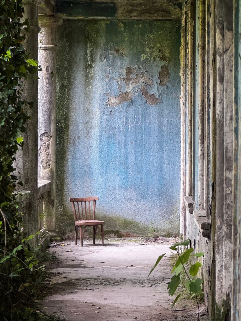 Verlassener, von Pflanzen überwucherter Gang mit verwitterter blauer Wand und einem einzelnen Holzstuhl