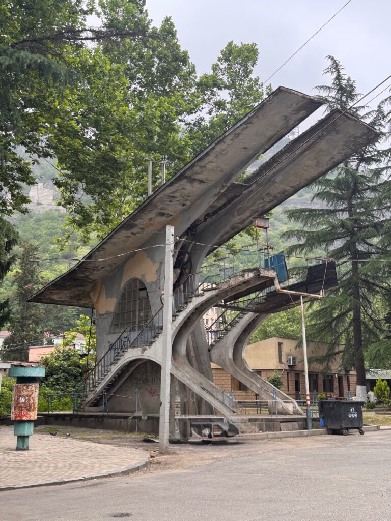 Modernistische Seilbahnstation mit zwei schräg nach oben verlaufenden Dachteilen und Treppenaufgängen mit Bäumen im Hintergrund