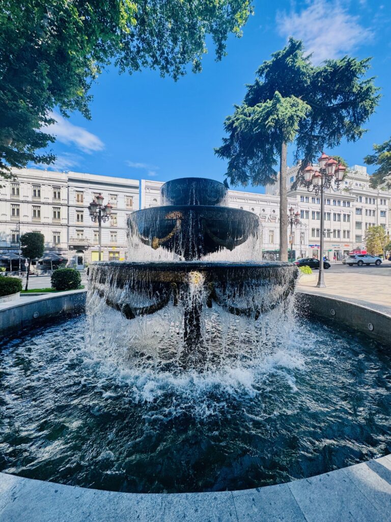 Mehrstufiger runder Brunnen mit Wasserfall in einem städtischen Park vor einem historischen Gebäude