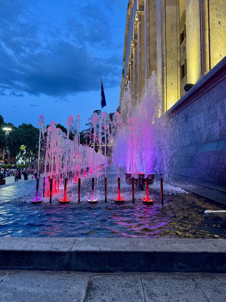 Springbrunnen mit rot und violett beleuchtetem Wasser vor beleuchtete Parlamentsgebäude in Tiflis, Georgien bei Abenddämmerung