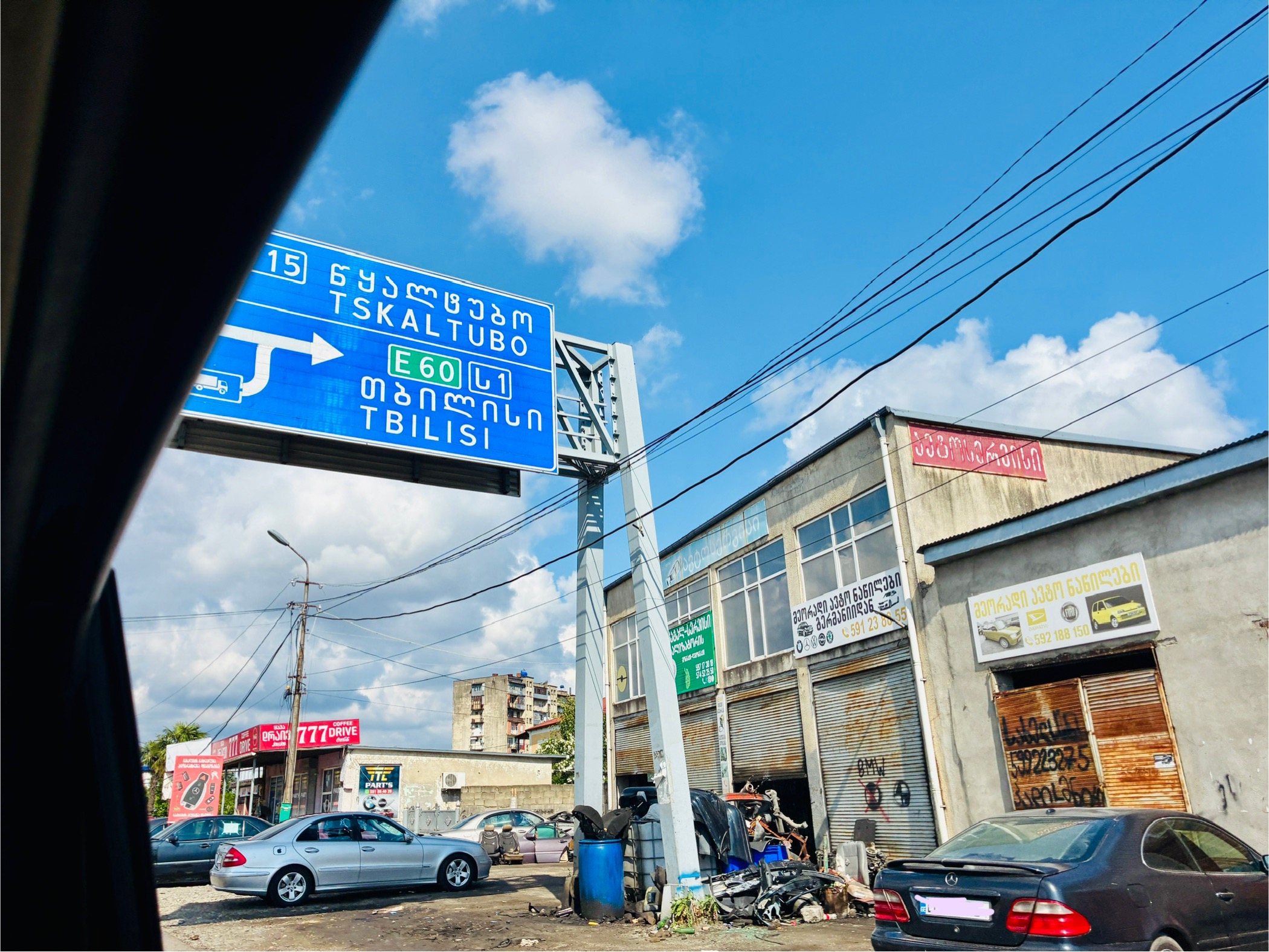 Straßenszene mit blauem Verkehrsschild in georgischer und lateinischer Schrift, das auf Tskaltubo und Tiflis verweist, umgeben von Gebäuden, parkenden Autos und einem klaren Himmel mit Wolken