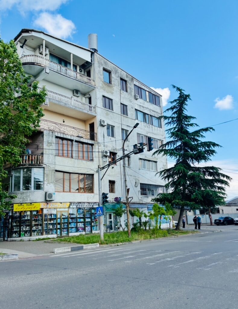 Mehrstöckiges Wohn- und Geschäftshaus mit Balkon, darunter ein Laden mit Schaufenstern und gelbem Schild, an einer Straße mit Fußgängerüberweg und zwei Personen auf dem Gehwegm neben einem großen Nadelbaum, der Putz des Hauses bröckelt etwas ab
