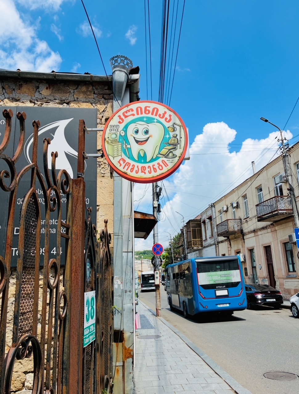 Rundes Schild mit lachendem Zahn und Zahnarztutensilien mit georgischen Schriftzeichen an einer Straßenlaterne in einer Stadtstraße mit Bus und niedrigen Gebäuden