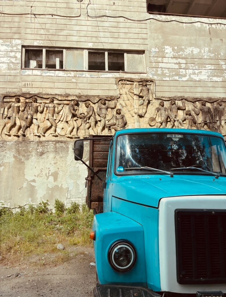 Altmodischer blauer Lastwagen vor einer Wand mit einem reliefartigen Wandbild aus Figuren in Bewegung