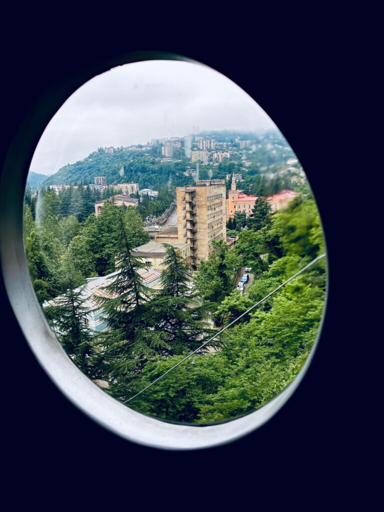 Blick durch Bullauge einer Seilbahnkabine auf bewaldete Hügel und mehrere Gebäude in einer Stadt