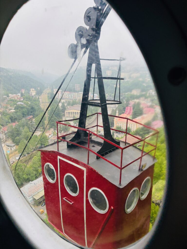 Rote Gondel einer Seilbahn mit runden Fenstern und Dachgeländer, aufgenommen durch das Bullauge der entgegenlaufenden Gondel