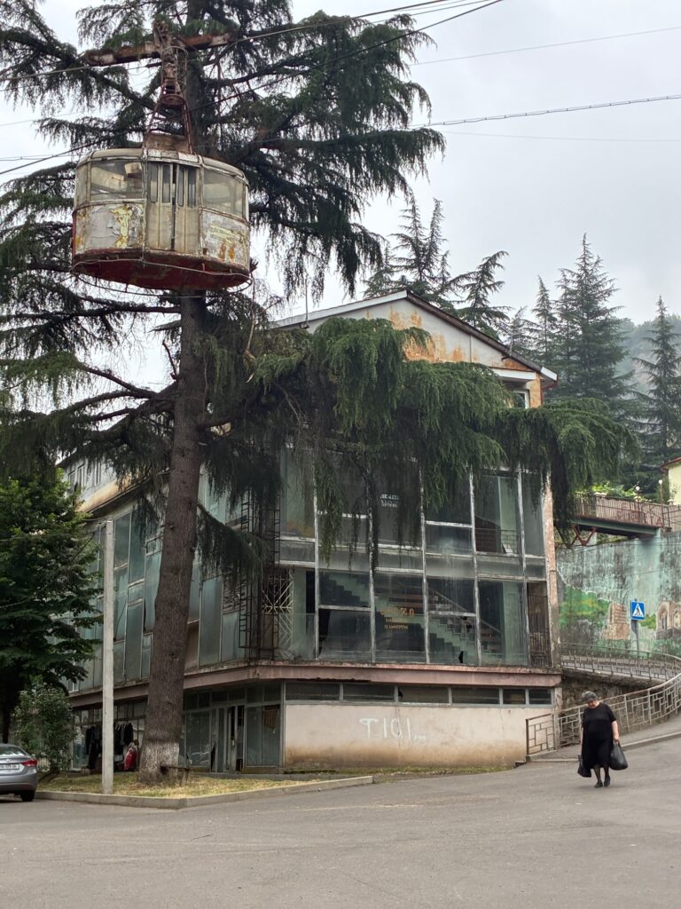 Verrostete Gondel einer Seilbahn hängt an einem Baum vor einem mehrstöckigen Gebäude mit großen Fenstern und eine Frau in schwarzen Kleidern, die auf der Straße gehen