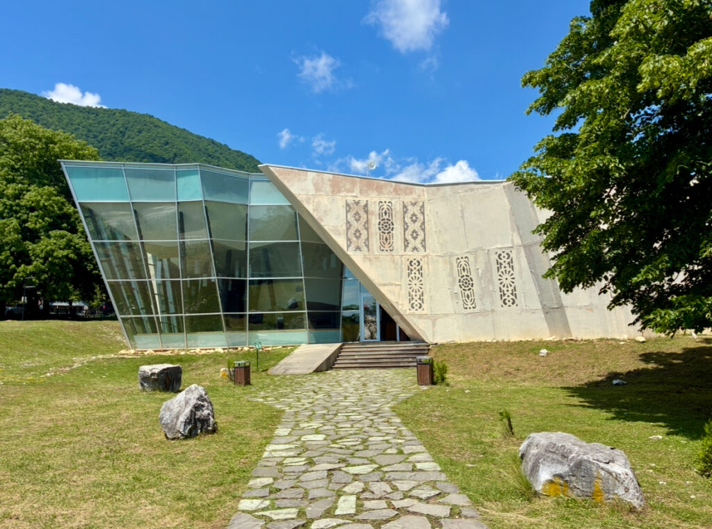 Modernes Gebäude mit schrägen Glas- und Betonflächen vor bewaldetem Hügel und blauem Himmel
