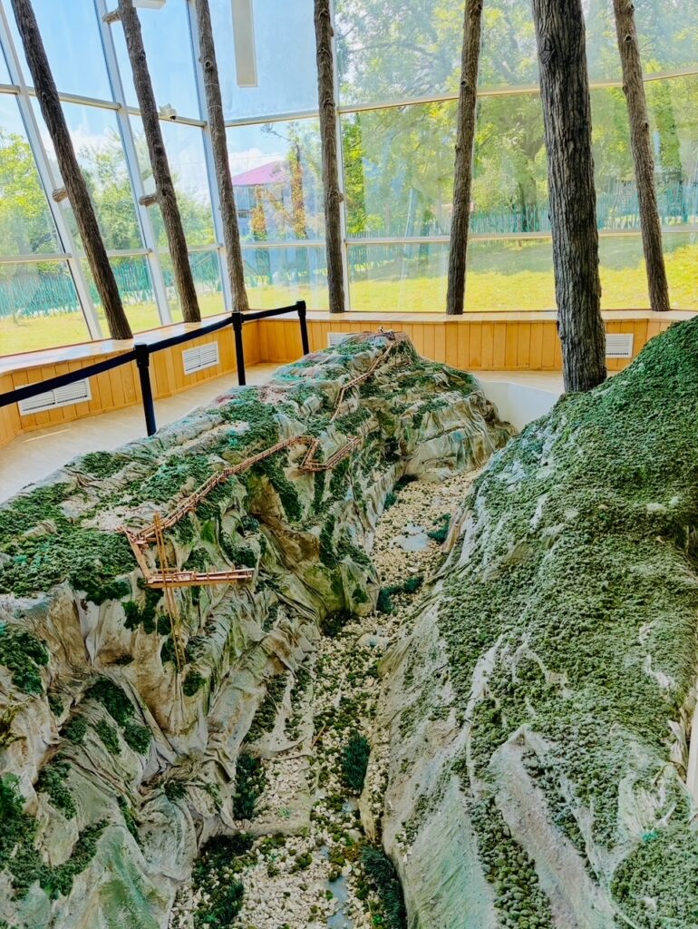 Modell einer bergigen Landschaft mit steilen Klippen und einem schmalen Pfad, umgeben von einem Raum mit großen Fenstern und Baumstämmen als Stützen