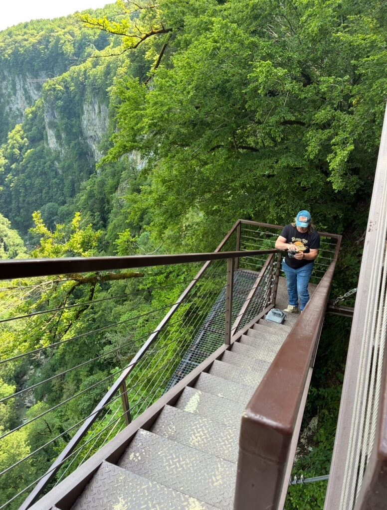 Person mit blauer Kappe und schwarzem T-Shirt steht auf einer Metalltreppe neben einem bewaldeten Felsenhang