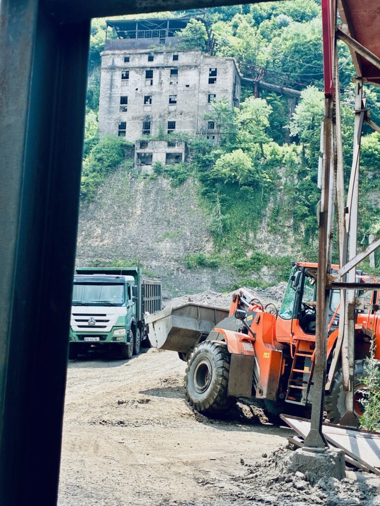 Baustelle mit orangefarbenem Radlader und weißem Lkw vor einem bewaldeten Hang mit altem, mehrstöckigem Gebäude