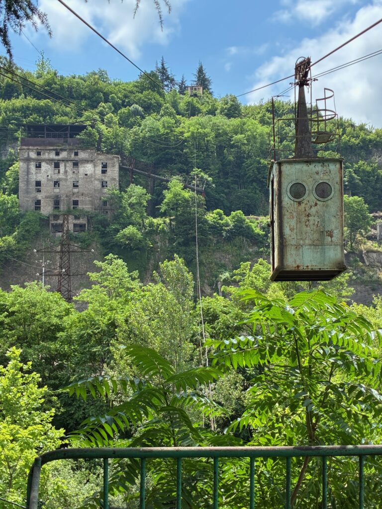 Verrostete, hängende Kabine einer Seilbahn vor bewaldetem Hügel mit altem, verlassenem Gebäude im Hintergrund