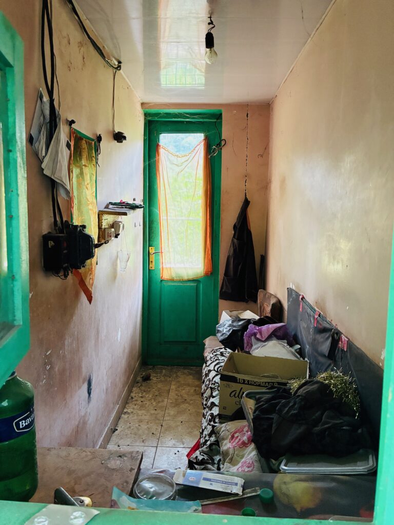Schmaler verlassener Raum mit grünem Türrahmen, einer Tür mit halbtransparentem Vorhang, einer Sitzbank mit Kartons und hingeworfener Kleidung sowie einer hängenden Jacke an der Wand