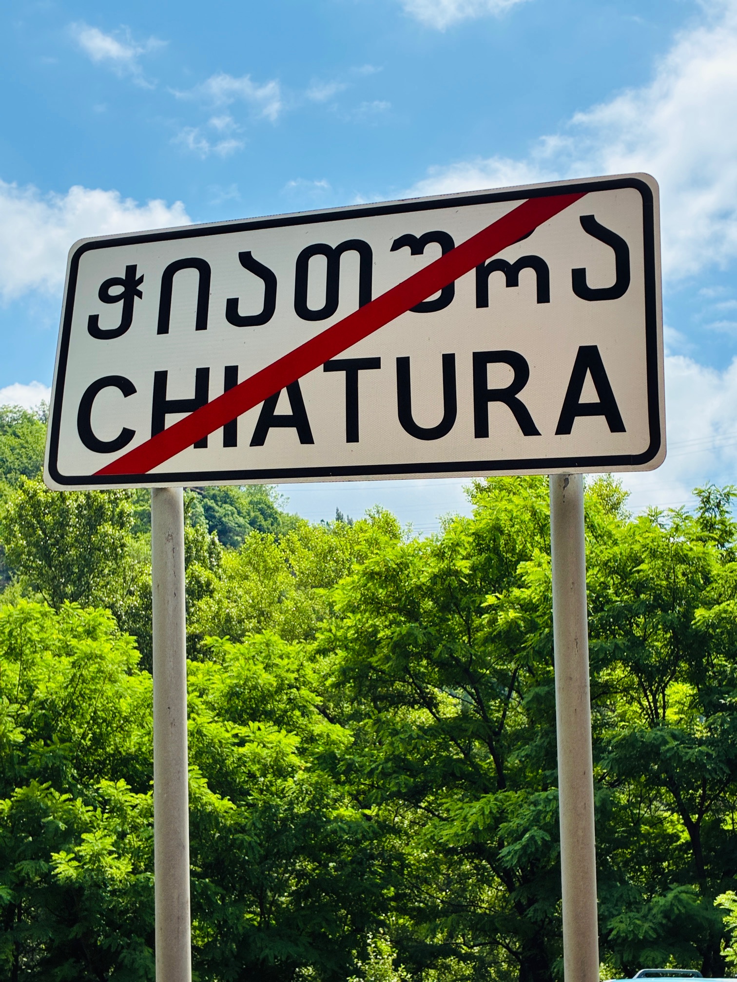 Ortsschild mit durchgestrichenem Schriftzug 'CHIATURA' in lateinischer und georgischer Schrift vor bewaldetem Hintergrund unter blauem Himmel mit freundlichen Wolken
