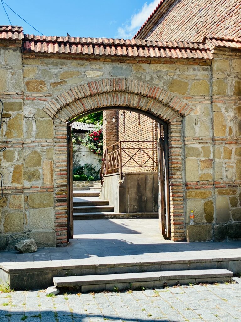Steinerner Torbogen mit Ziegelumrandung führt zu einer Treppe und einem Innenhof mit Mauer und Geländer