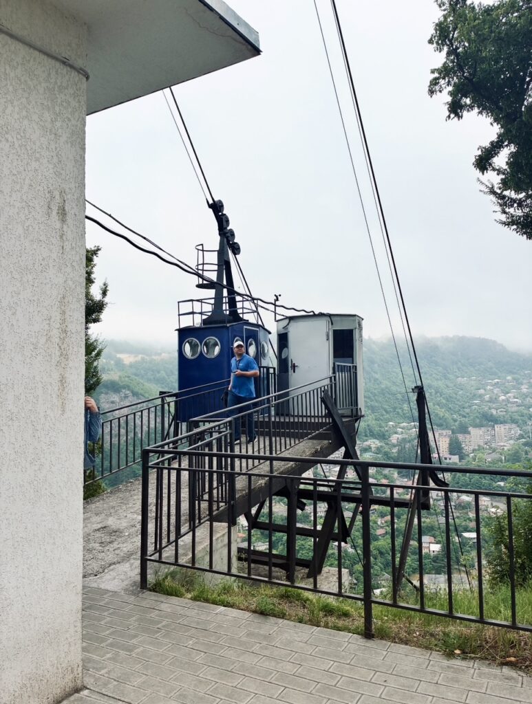 Bergstation einer Seilbahn mit blauem Kabinenaufbau und einer Person in blauem T-Shirt auf einer Plattform mit Geländer