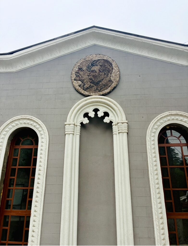 Fassade eines Gebäudes mit zwei hohen, schmalen Fenstern und einem mittigen, verzierten Bogen sowie einem runden Mosaik mit den Profilen von Lenin und Stalin darüber