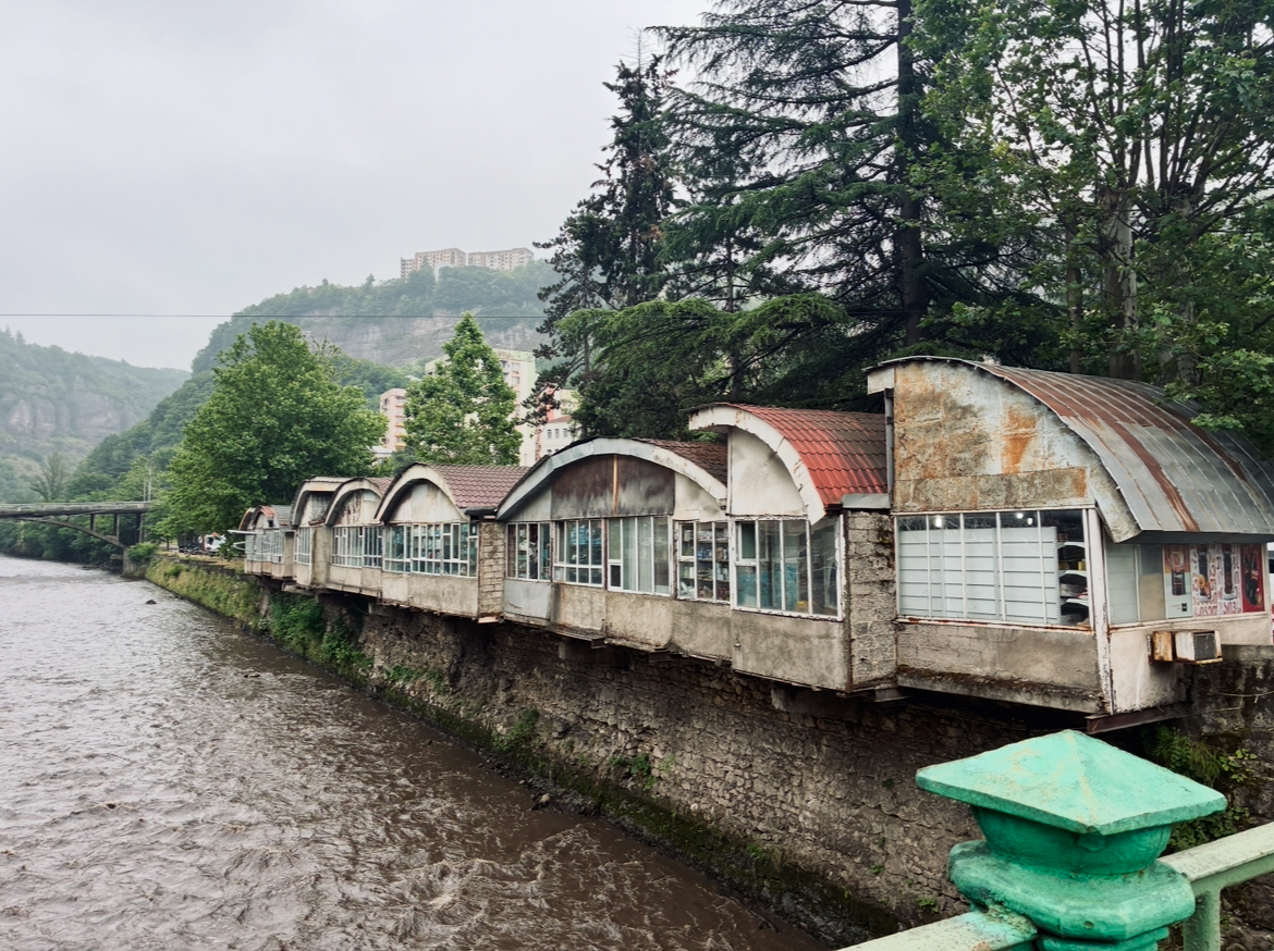 Reihe von verrottenden, halbrunden, fensterreichen Gebäuden auf einer Steinmauer am Flussufer mit bewaldeten Hügeln im Hintergrund