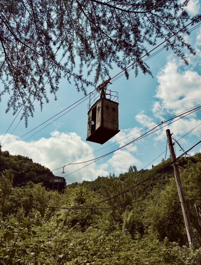 Verrostete, verlassene Seilbahn-Gondel hängt am Seil über bewaldetem Hügel unter blauem Himmel mit Wolken