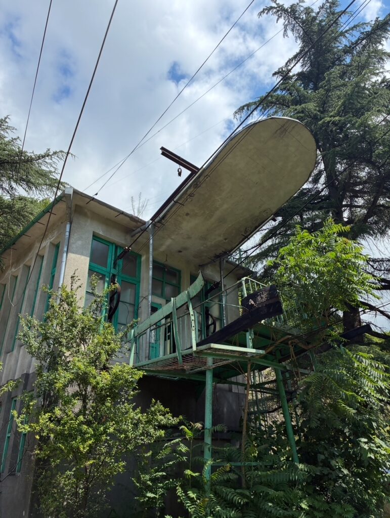 Verfallene Talstation einer Seilbahn mit grünem Holzgerüst und großem, ovalem Sonnenschutzdach über einem Balkon, umgeben von Bäumen