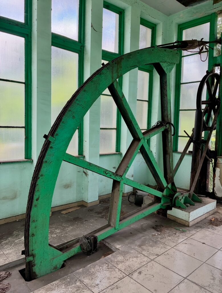 Großes grünes Antriebsrad einer Seilbahn mit Speichen in einem Raum mit hohen Fenstern