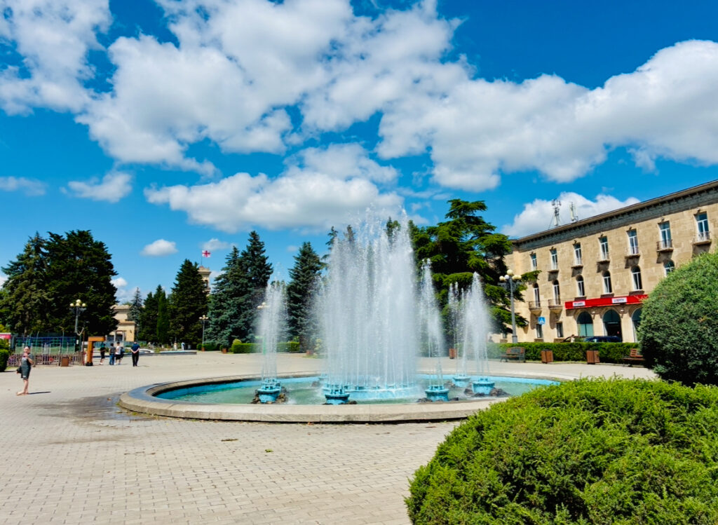 Springbrunnen mit mehreren Wasserstrahlen auf gepflastertem Platz vor einem mehrstöckigen Gebäude und Büschen unter blauem Himmel mit Wolken