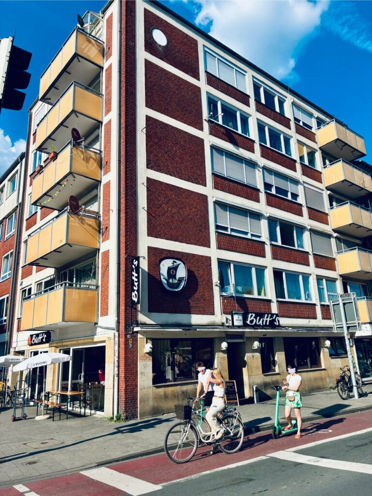 Mehrstöckiges Midcentury Wohn- und Geschäftshaus mit roter Backsteinfassade und weißen Fensterrahmen an einer Straßenecke mit Fahrradfahrern und Fußgängern unter blauem Himmel mit freundlichen Wolken