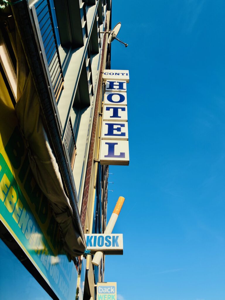 Von unten nach oben fotografierte Fassade mit vertikalem Schild 'HOTEL' und darunter kleinem Schild 'KIOSK' vor blauem Himmel, quer darüber eine stilisierte Zigarette