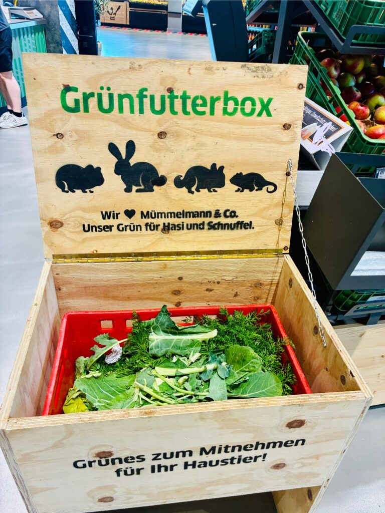 Offene Holzkiste mit frischem Grünfutter und Aufschrift 'Grünfutterbox' sowie Silhouetten von Kleintieren auf dem Deckel