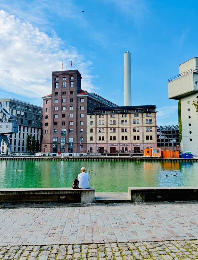Person sitzt auf einer Mauer am Wasser vor mehreren mehrstöckigen Gebäuden und einem hohen Schornstein