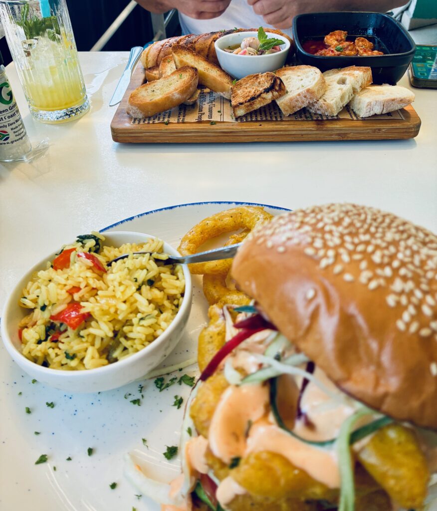 Teller mit Burger und buntem Reis mit Gemüse, im Hintergrund Holzbrett mit getoastetem Brot