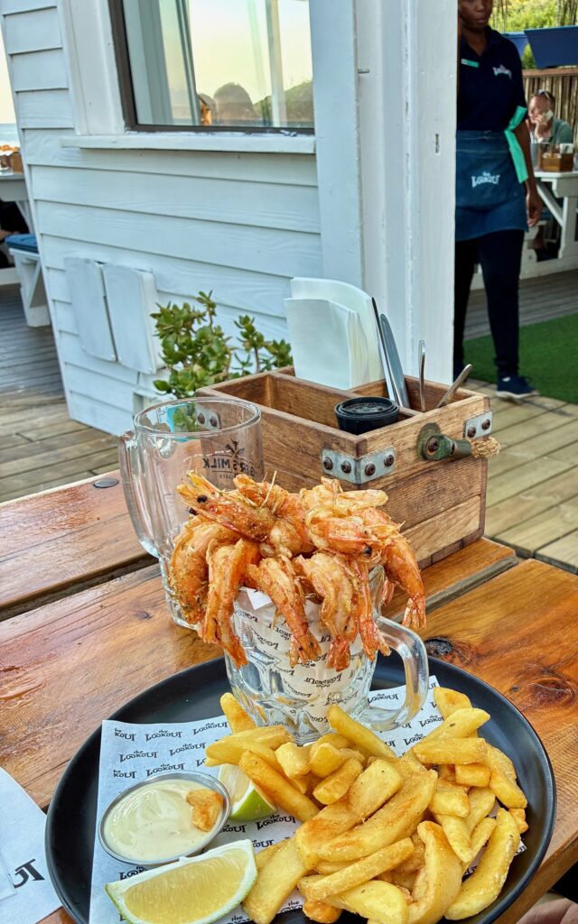 Teller mit Pommes Frittes und einem Limettenschnitz, dazu ein Glas mit frischen Shrimps, im Hintergrund ein Holzkasten mit Besteck