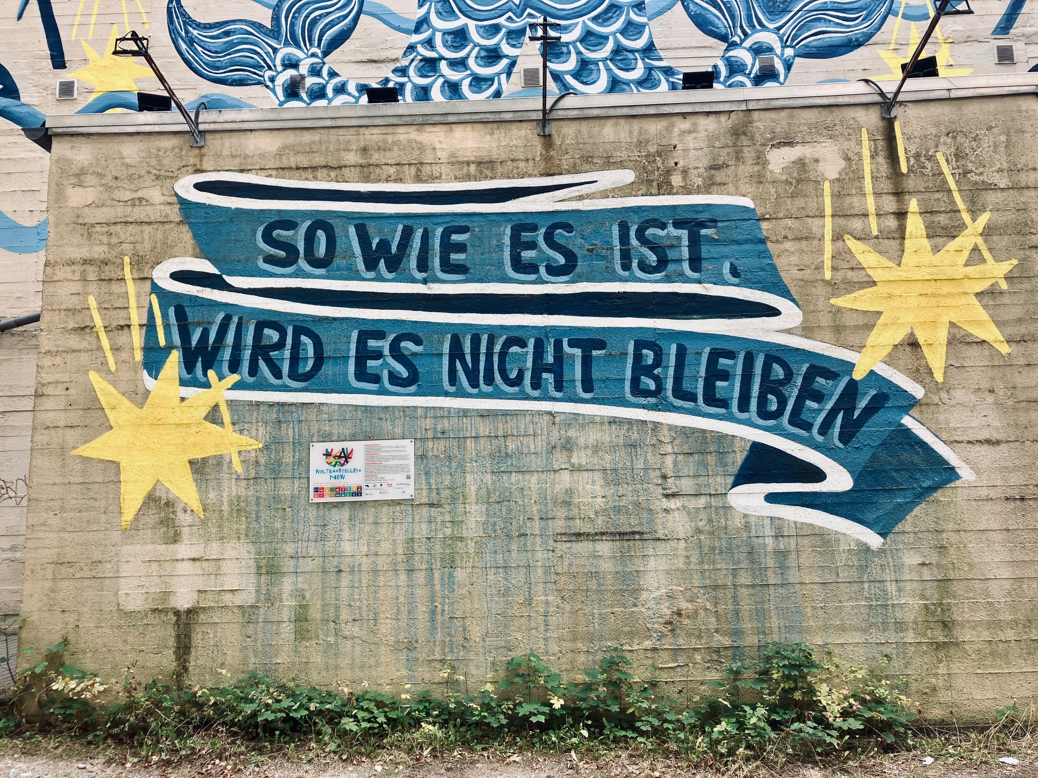 Graffiti an einer Betonwand mit der Schrift 'SO WIE ES IST, WIRD ES NICHT BLEIBEN' auf einem blauen Band zwischen zwei gelben Sternen
