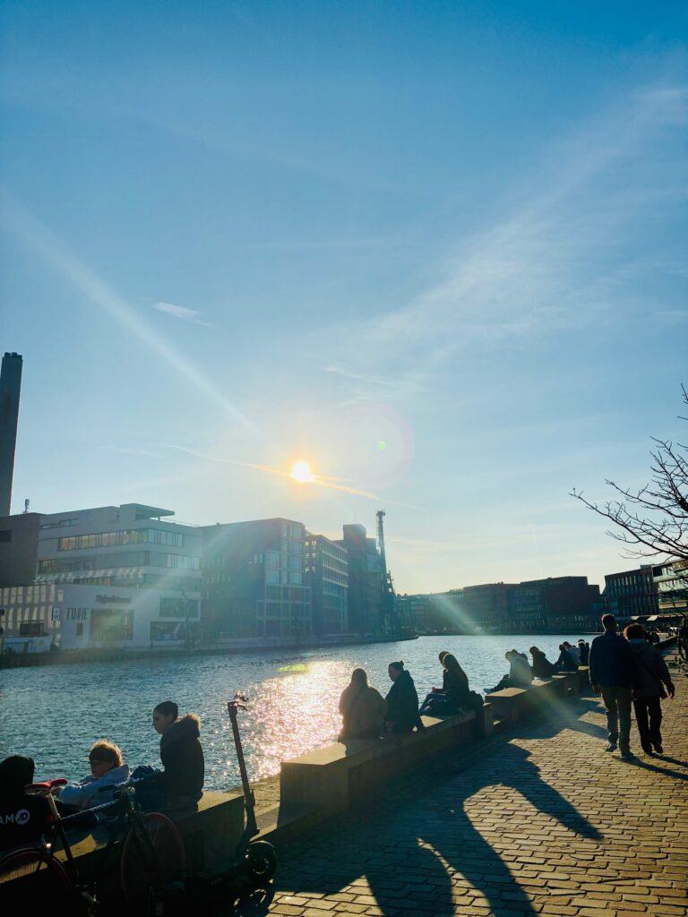 Menschen sitzen und stehen am sonnigen Ufer am Hafen von Münster mit moderner Stadtbebauung im Hintergrund