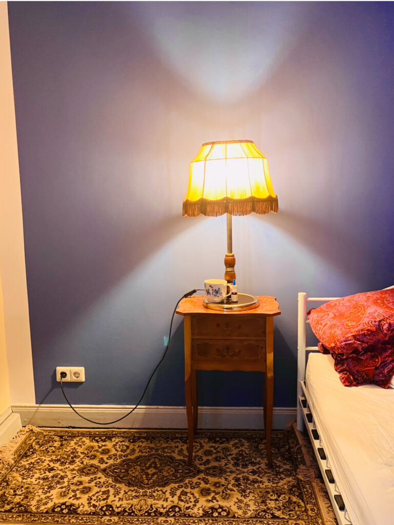 Beistelltisch mit Tischlampe und Tasse neben einem Bett mit buntem Kissen vor blauem Wandhintergrund, davor gemusterter Teppich