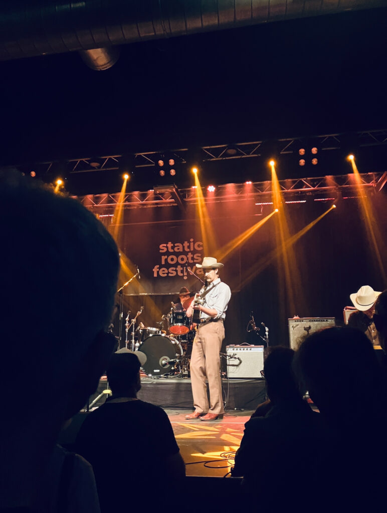 Der Musiker Todd Day Wait mit Cowboyhut spielt Gitarre auf Bühne mit Beleuchtung und Schriftzug 'static roots festival' im Hintergrund