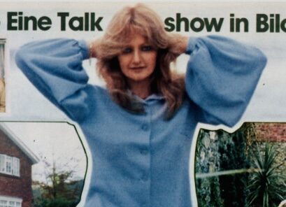 Frau mit langen, gewellten Haaren trägt einen blauen Pullover und hält die Hände an den Kopf vor einem Banner mit teilweise sichtbarem Text. Die Frau ist die Sängerin Bonnie Tyler auf einem Foto von 1978