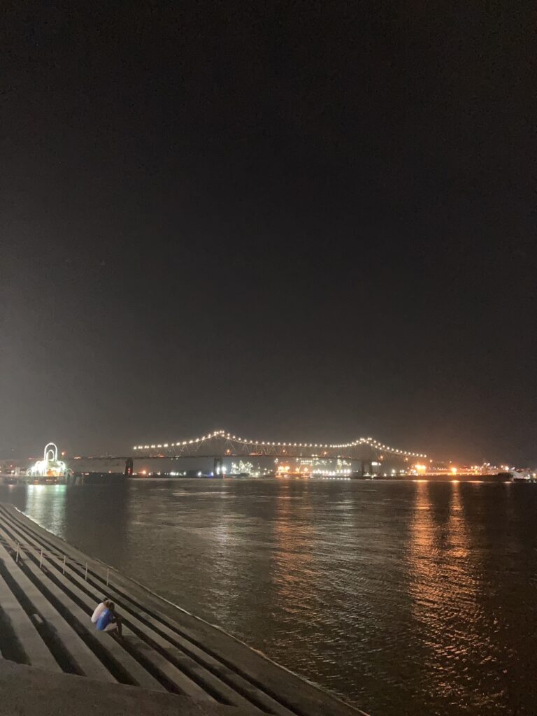 Blick auf eine beleuchtete Hängebrücke über einen Fluss bei Nacht, im Vordergrund Treppenstufen und eine Person, die auf den Stufen sitzt