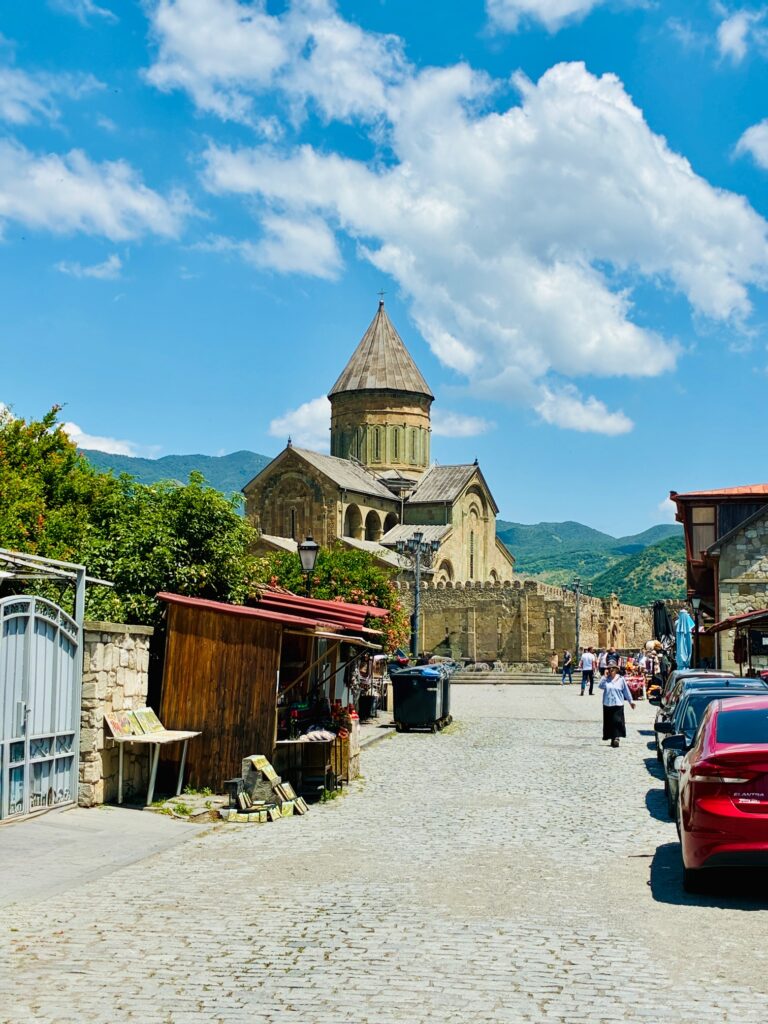 Kopfsteinpflasterstraße mit Marktständen und parkenden Autos führt zu einer historischen Kirche mit rundem Turm vor bewaldeten Bergen unter blauem Himmel mit Wolken