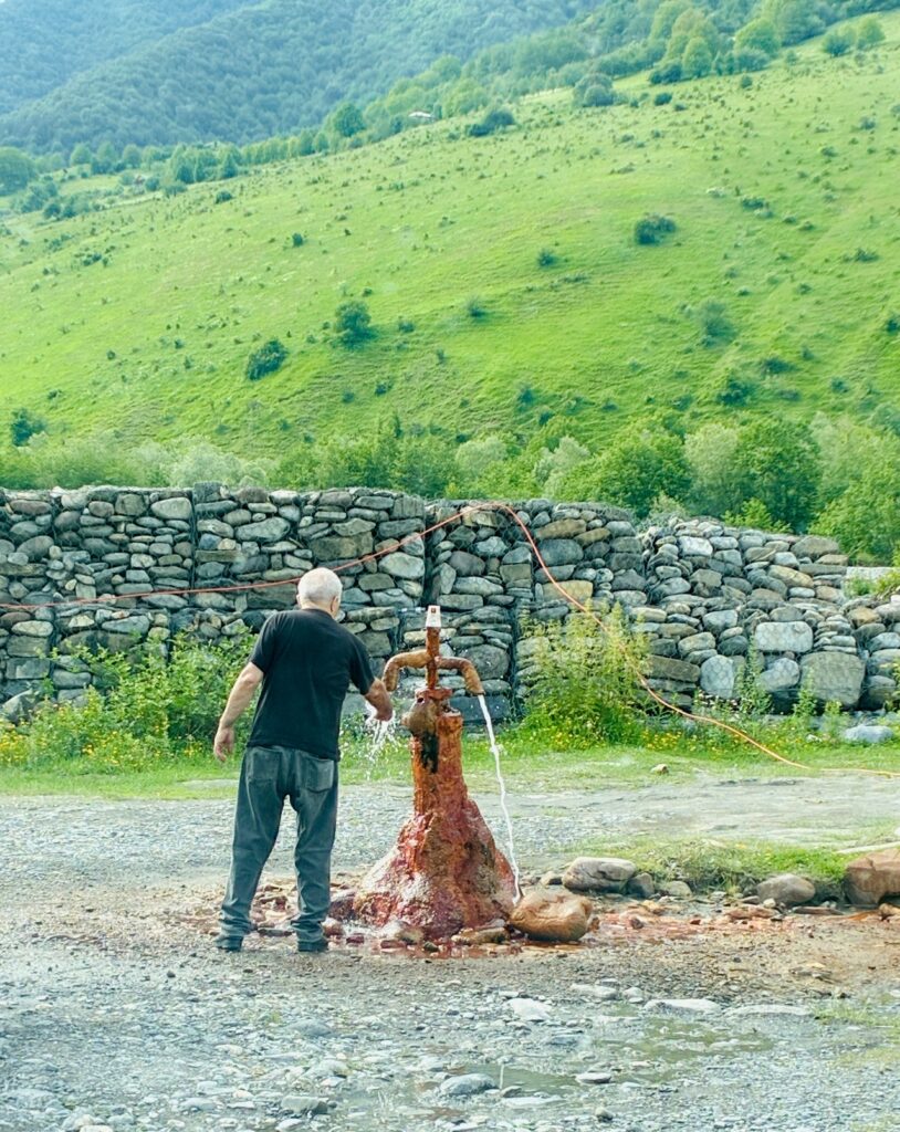 Ein Mann steht an einem oxiederten alten Quellbrunnen aus Metall, aus dem Wasser fließt, vor einer niedrigen Steinmauer und einem bewaldeten Hügel im Hintergrund
