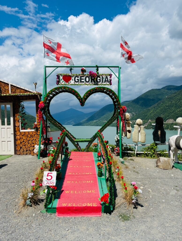 Herzförmiger Blumenbogen mit dem Schriftzug 'I love GEORGIA' über einem roten Teppich mit dem Wort 'WELCOME' mehrfach darauf, im Hintergrund Berge, ein türkisfarbener See und zwei wehende georgische Flaggen