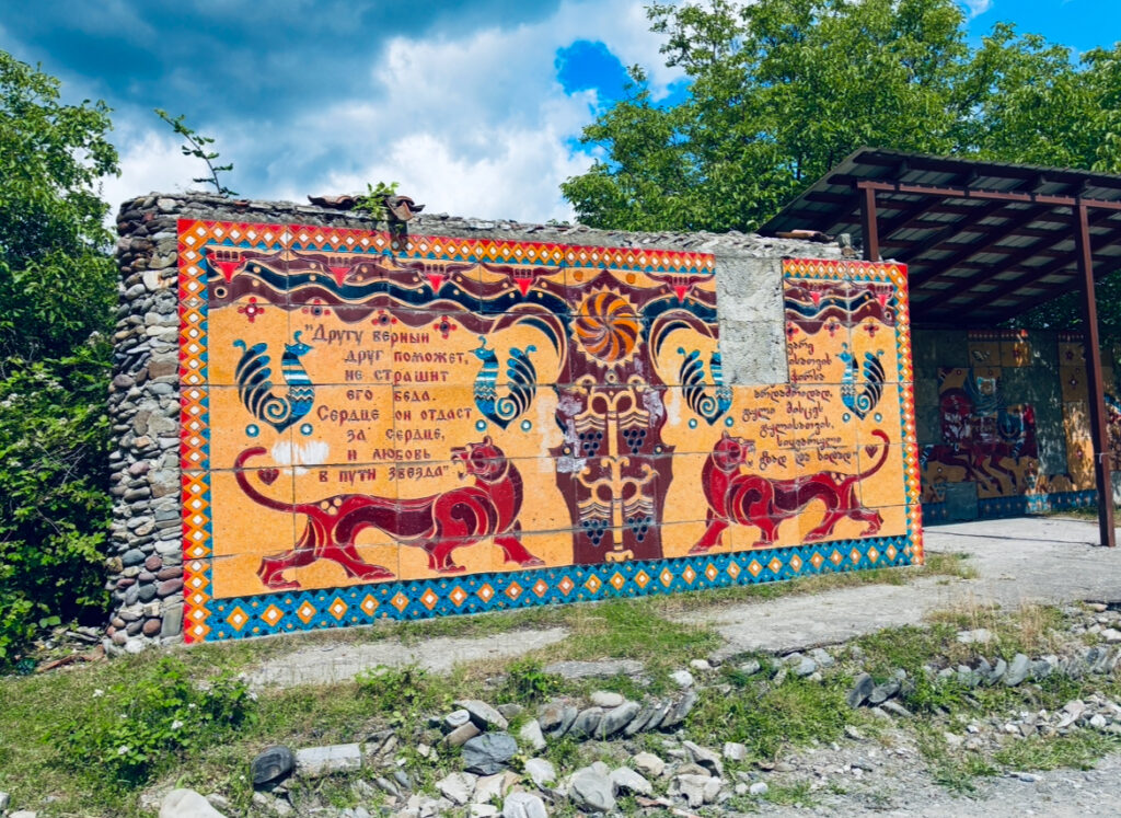 Großes Wandmosaik mit zwei stilisierten Löwen, dekorativen Mustern und kyrillischem Text, umgeben von Natur und einem überdachten Bereich
