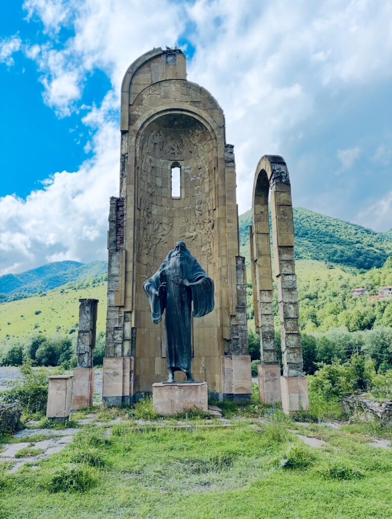 Statue einer stehenden Figur mit ausgebreiteten Armen vor einer hohen, bogenförmigen Steinruine in einer grünen Landschaft mit Bergen im Hintergrund