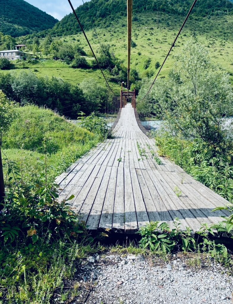 Hängende Holzbrücke mit Seilgeländern über einem Fluss in einer grünen Berglandschaft