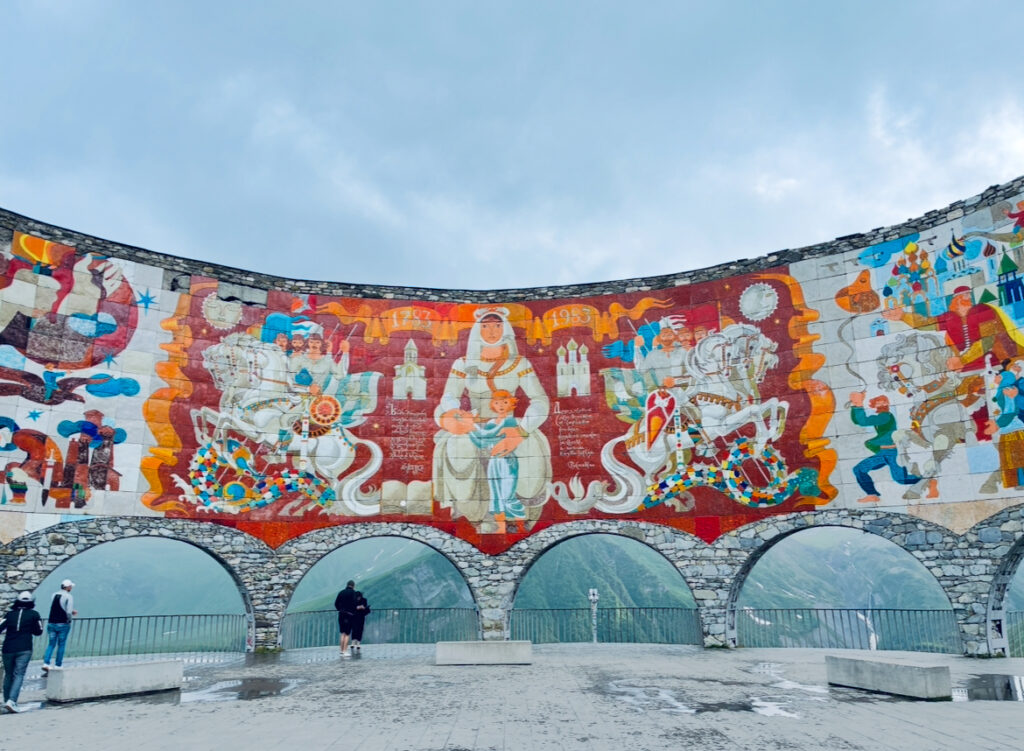 Großes, farbenfrohes Wandgemälde mit verschiedenen Figuren und Symbolen auf einer halbkreisförmigen Steinmauer mit Bögen, davor stehen mehrere Personen