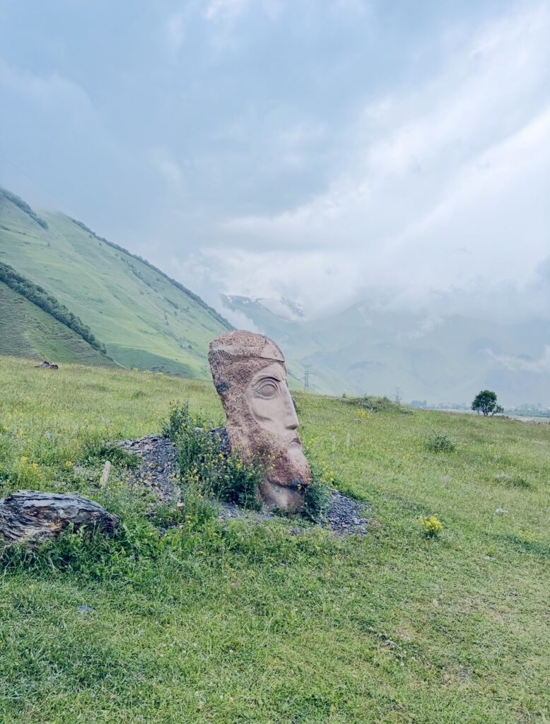 Große steinerne Kopfskulptur auf einer grünen Wiese vor bewölktem Himmel und Bergen im Hintergrund
