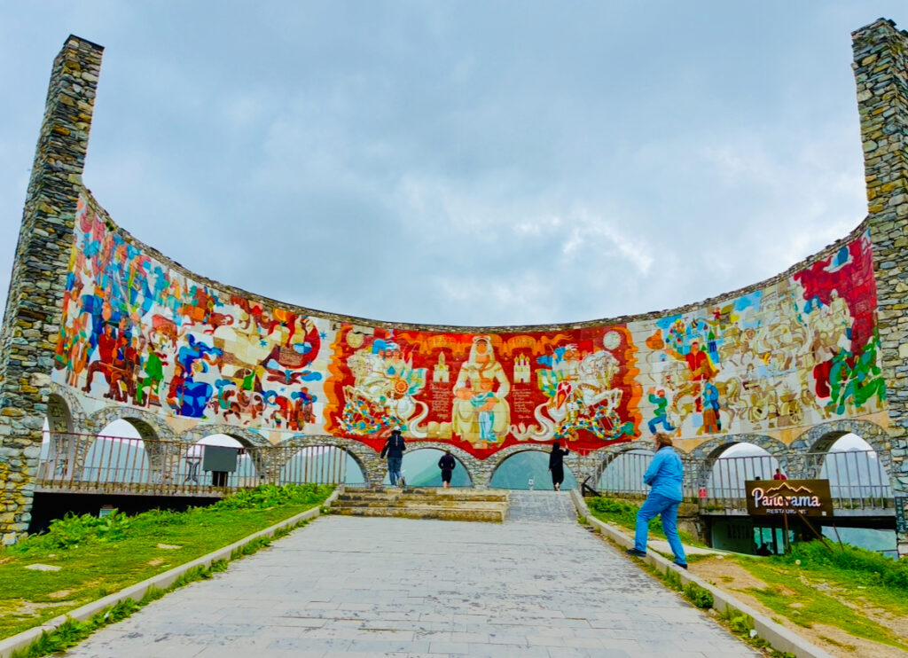 Bunte Wandmalerei auf halbkreisförmiger Mauer mit mehreren Personen davor und Schild mit Schriftzug 'Panorama'