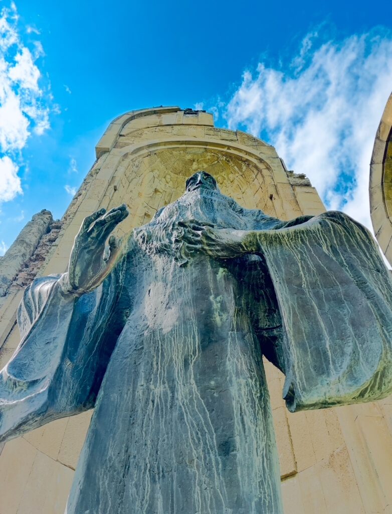 Von unten aufgenommene Statue mit ausgebreiteten Armen und gespreizten Händen vor einem hellen Himmel und einem steinernen Bauwerk